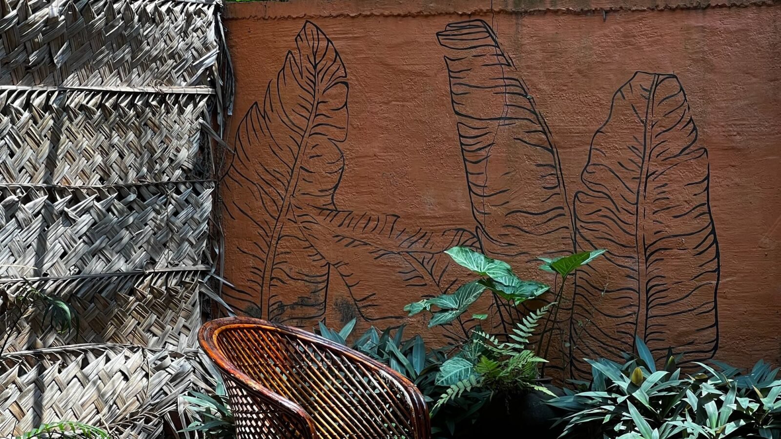 Compact outdoor patio with wicker chair, small table, fence, and tropical plants.